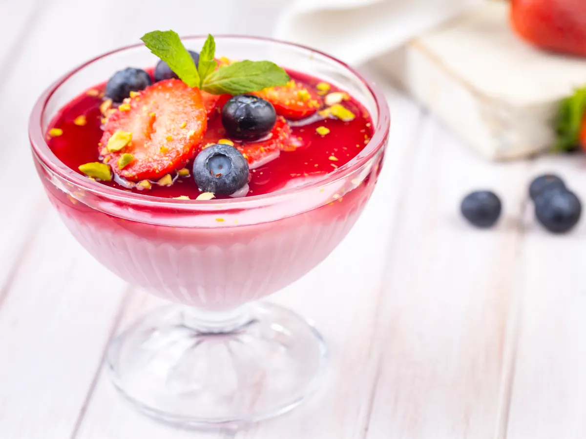 Una copa de cristal con mousse rosa, cubierta con fresa laminada, arándanos y una hoja de menta.