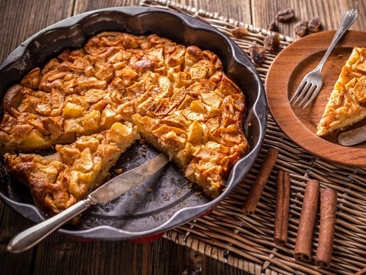 Una tarta de manzana recién horneada en su molde, con una porción servida en un plato de madera y ramas de canela al lado.